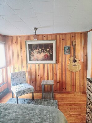 Una habitación con paneles de madera, un cuadro enmarcado, una guitarra, una silla y una mesita.