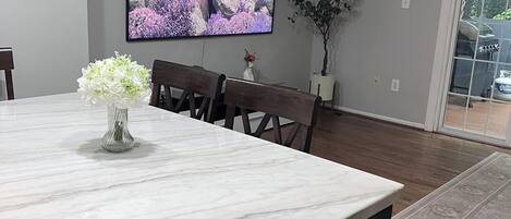 A dining area with a marble table, wooden chairs, a flat-screen TV on the wall, and a potted plant.