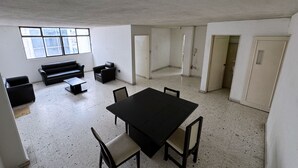 Una habitación amplia con una gran mesa negra y cuatro sillas, un sofá negro y una mesita auxiliar negra. Dos ventanas aportan luz natural.