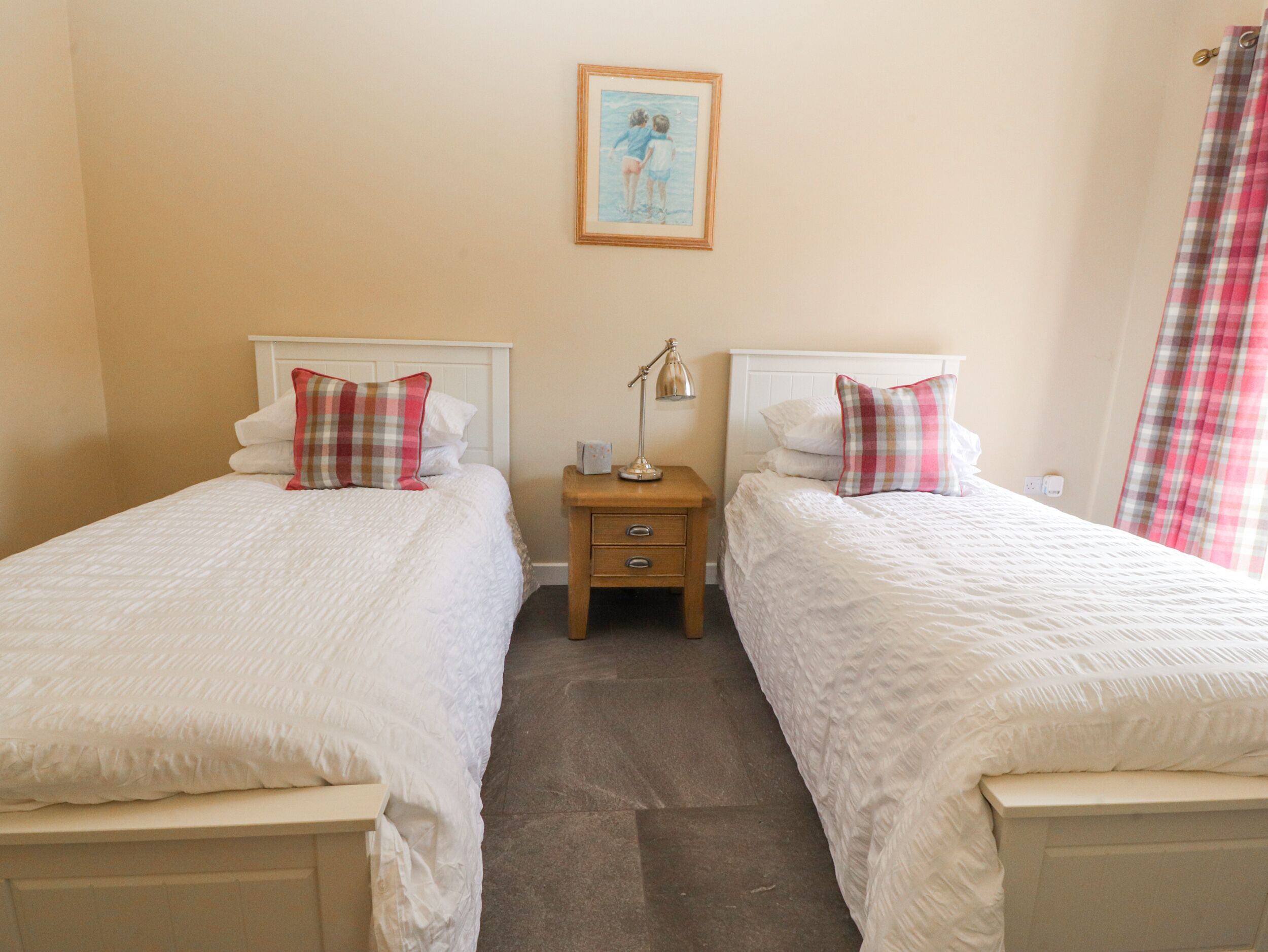 A hotel room with two single beds, a nightstand with a lamp, a framed picture on the wall, and a window with a checkered curtain.