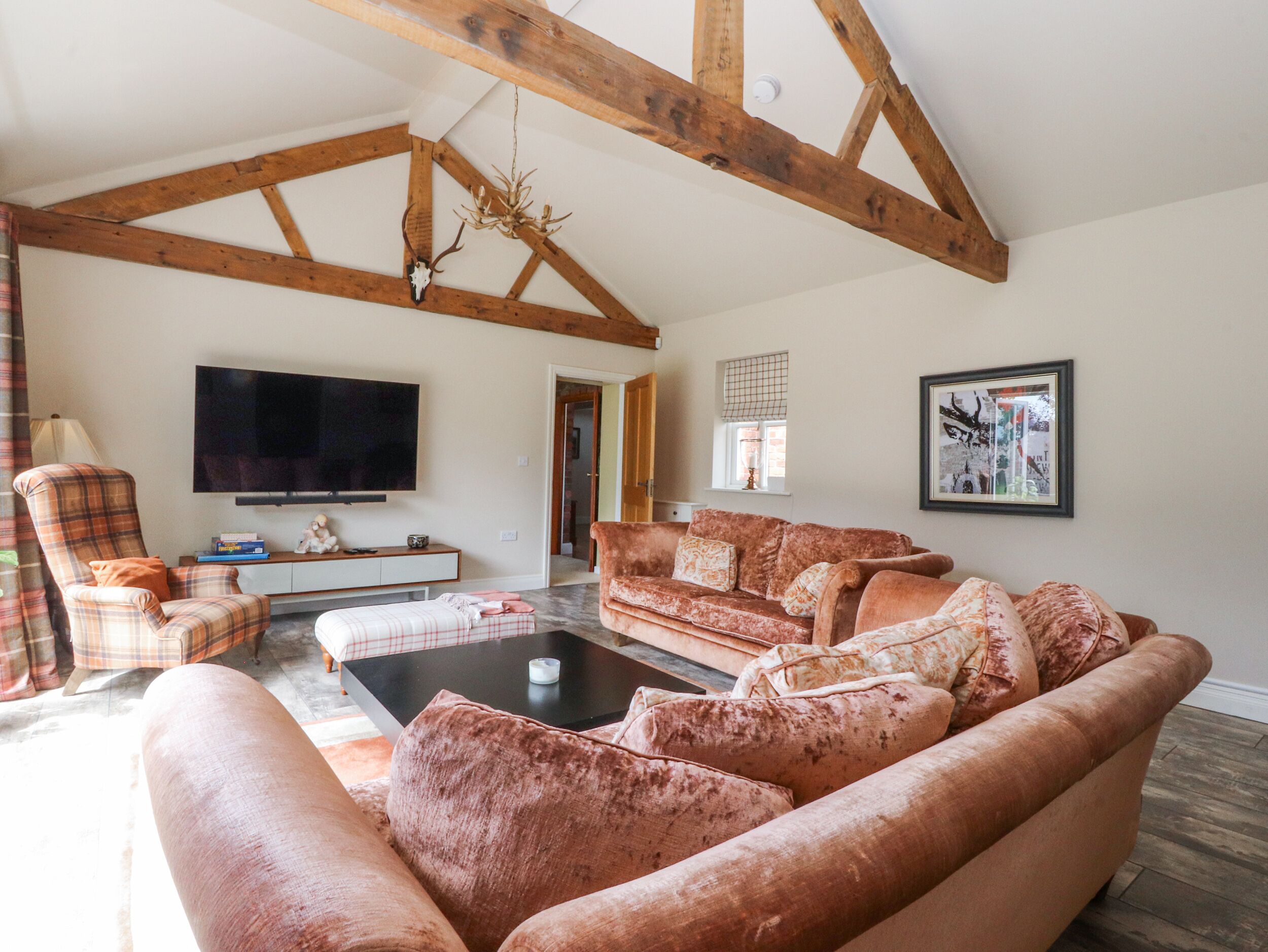 A spacious living room with a high ceiling, wooden beams, a large flat-screen TV, a fireplace, and a cozy seating area with a patterned sofa and armchairs.