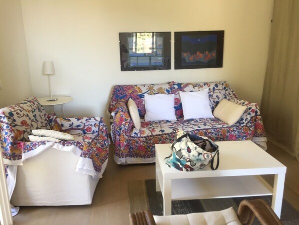 Sala de estar com dois sofás, uma mesa pequena e uma luminária. Há duas imagens emolduradas na parede.