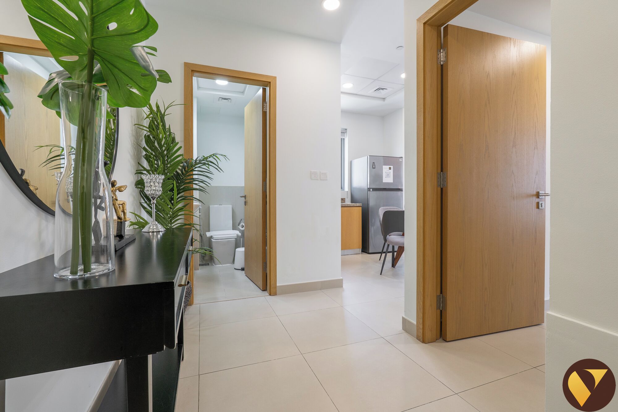 A hallway with a black console table, a mirror, and a potted plant. There is a wooden door on the right and a white door in the background leading to a kitchen area.