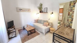 Un espacio habitable pequeño con un sofá, una mesita, un televisor en un estante y una estantería con una planta.