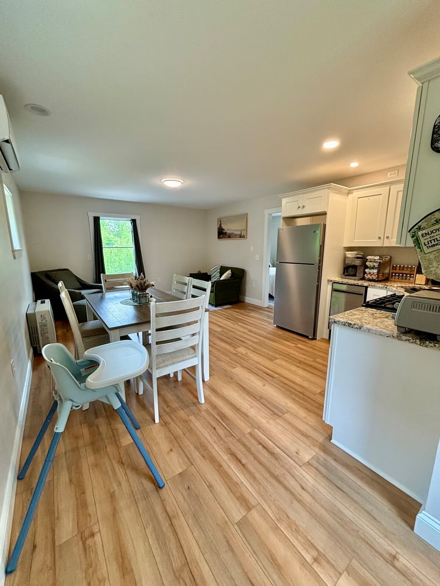A modern kitchen with a dining area, wooden flooring, and a view of greenery outside.