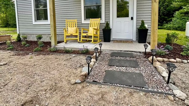 A house with a covered porch, two chairs, a door, and a small garden area. - Brand new 2BR, 1.5BA-close to amenities yet feels secluded. 2 story, AC, wifi! (Bridgton)