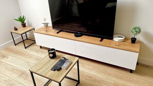 Ein modernes Wohnzimmer mit einem Flachbildfernseher auf einer Holzkonsole, einem Couchtisch mit Fernbedienung und zwei Beistelltischen mit Zierpflanzen.