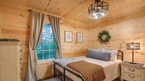 Een knus houten vertrek met een bed, een nachtkastje, een raam met gordijnen en ingelijste foto’s aan de muur.