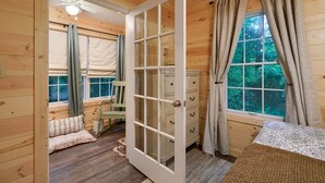 A wooden cabin interior with a large open door, a window with curtains, a chair, and a small table.