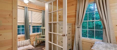Ein Holzblockhaus-Interieur mit einer großen offenen Tür, einem Fenster mit Vorhängen, einem Sessel und einem kleinen Tisch.