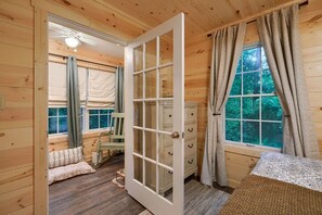 Interior de una cabaña de madera con una puerta grande entreabierta, una ventana con cortinas, una silla y una mesita.