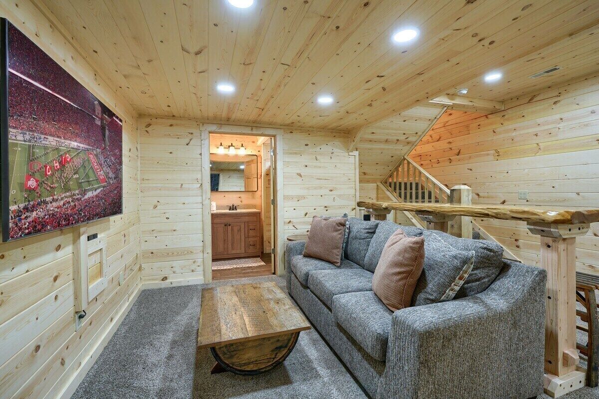 A cozy log cabin interior with a wooden ceiling, a gray sofa with cushions, a wooden coffee table, and a kitchen area visible through an open doorway.