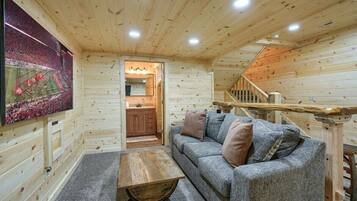 A cozy log cabin interior with a wooden ceiling, a gray sofa with cushions, a wooden coffee table, and a kitchen area visible through an open doorway.