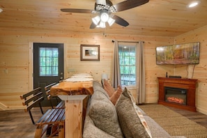 A cozy cabin interior with a wooden ceiling, a ceiling fan, a fireplace, a television, and a bench.
