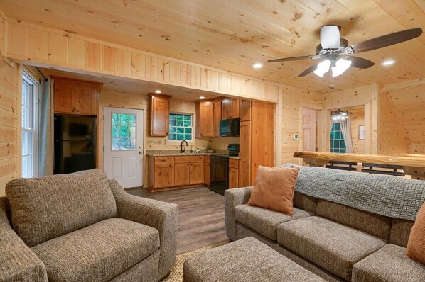 A cozy cabin interior with wooden walls, a kitchenette, a dining area, and two sofas.