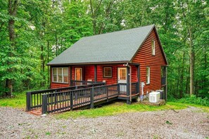 A cabin with a deck and a covered porch, surrounded by dense forest.