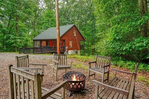 A cabin with a fire pit and wooden chairs in a forest setting.
