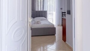 A hotel room with a bed, a desk, a chair, a window with curtains, and an air conditioning unit. - Villa Nomia (Ksamil)