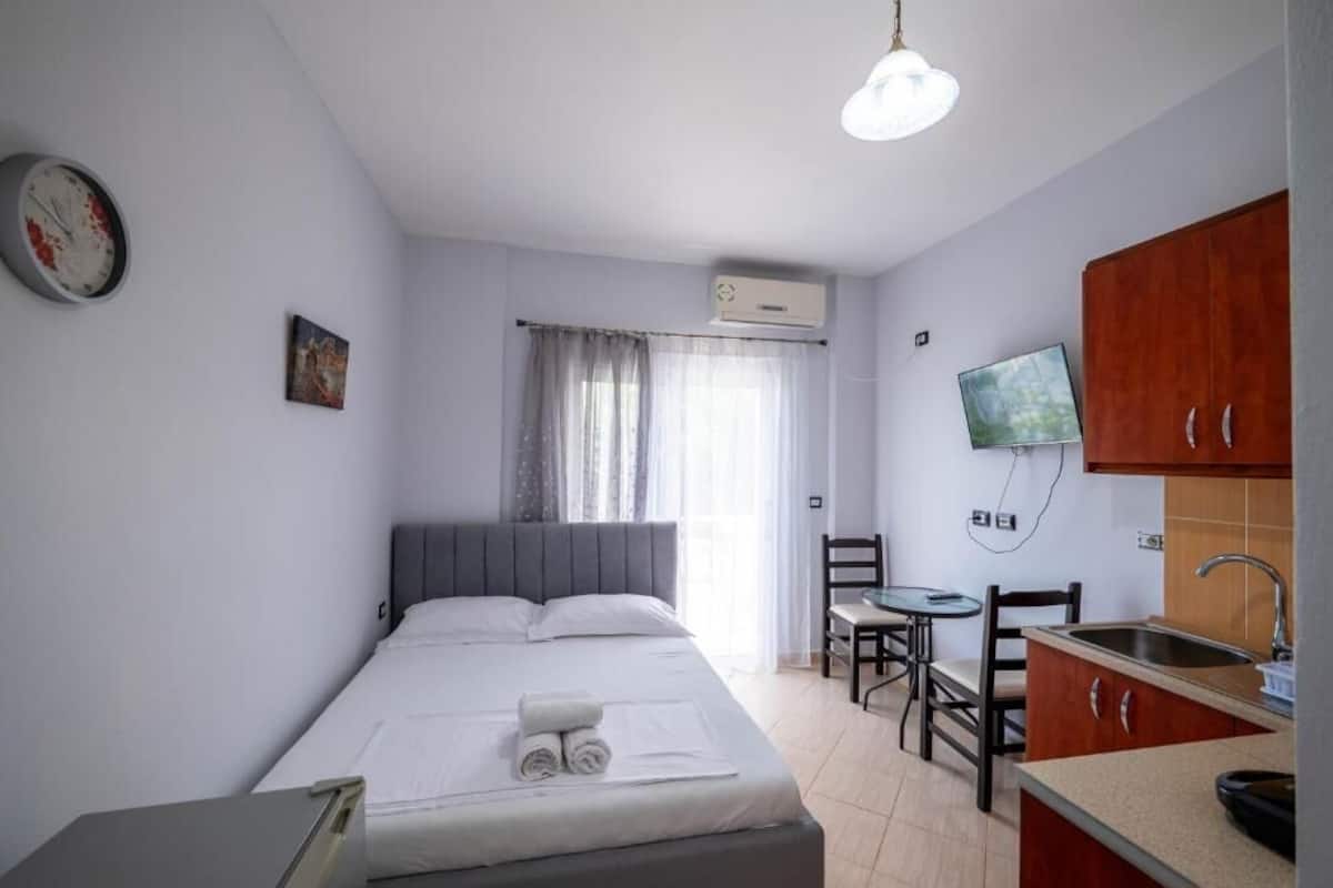 A compact hotel room with a bed, a small desk, a chair, a television, and a kitchenette.