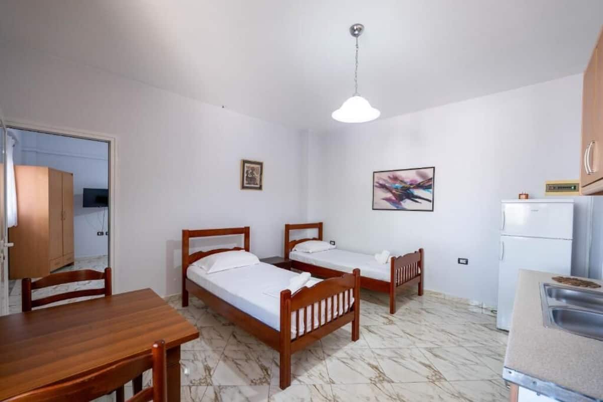 A room with two wooden beds, a wooden table, a small kitchenette, and a single light fixture.