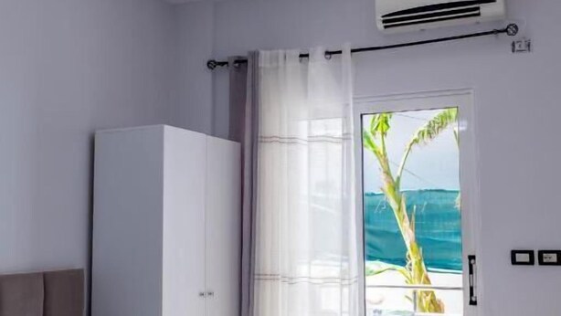 A hotel room with a bed, a wardrobe, an air conditioning unit, a curtain, and a view of the ocean. - Villa Nomia (Ksamil)