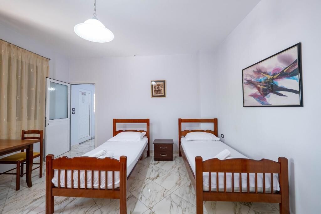 A room with two wooden beds, a dining table, a chair, a small bedside table, a framed picture on the wall, and a door.
