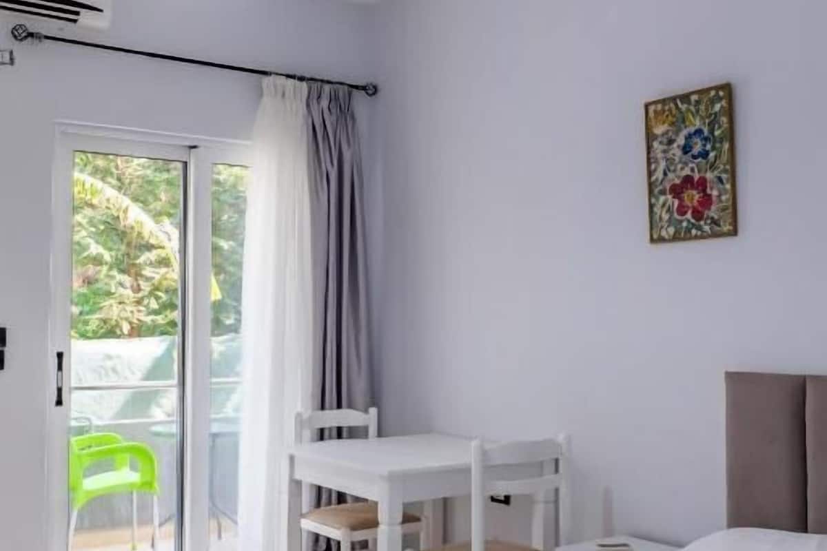A hotel room with a bed, a desk, a chair, a window with curtains, and a painting on the wall.