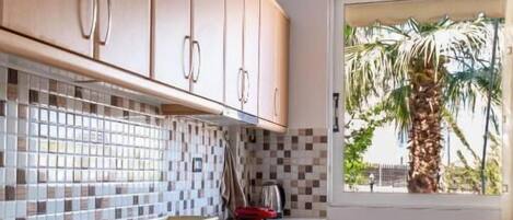 Eine Küche mit Holzausstattung, einer gefliesten Spritzschutzwand, einem Fenster mit Blick auf Palmen und einem Stuhl.