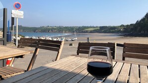 A wooden table with a glass of red wine and a bench overlooking a beach with boats and a no parking sign.