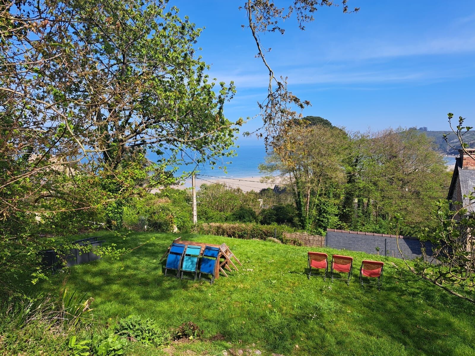 A grassy outdoor area with a view of the sea, a wooden structure with blue and red chairs, and a stone wall in the background.