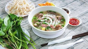 Eine Schüssel Suppe mit Nudeln, Fleisch und Gemüse, dazu frische Kräuter, Sojasprossen und verschiedene Dips auf einem Holztisch.