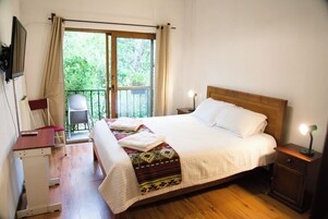 Habitación de hotel con una cama, lámparas de noche, un escritorio, una silla y vistas a zonas verdes por la ventana.