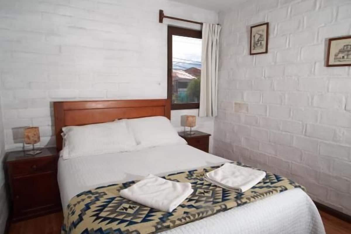 Un dormitorio con cama de madera, paredes de ladrillo blanco, ventana con cortinas e imágenes enmarcadas en la pared.