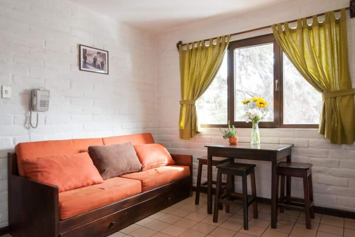 Una habitación acogedora con un sofá, una mesita y sillas. Se ve una ventana con cortinas y un cuadro en la pared.
