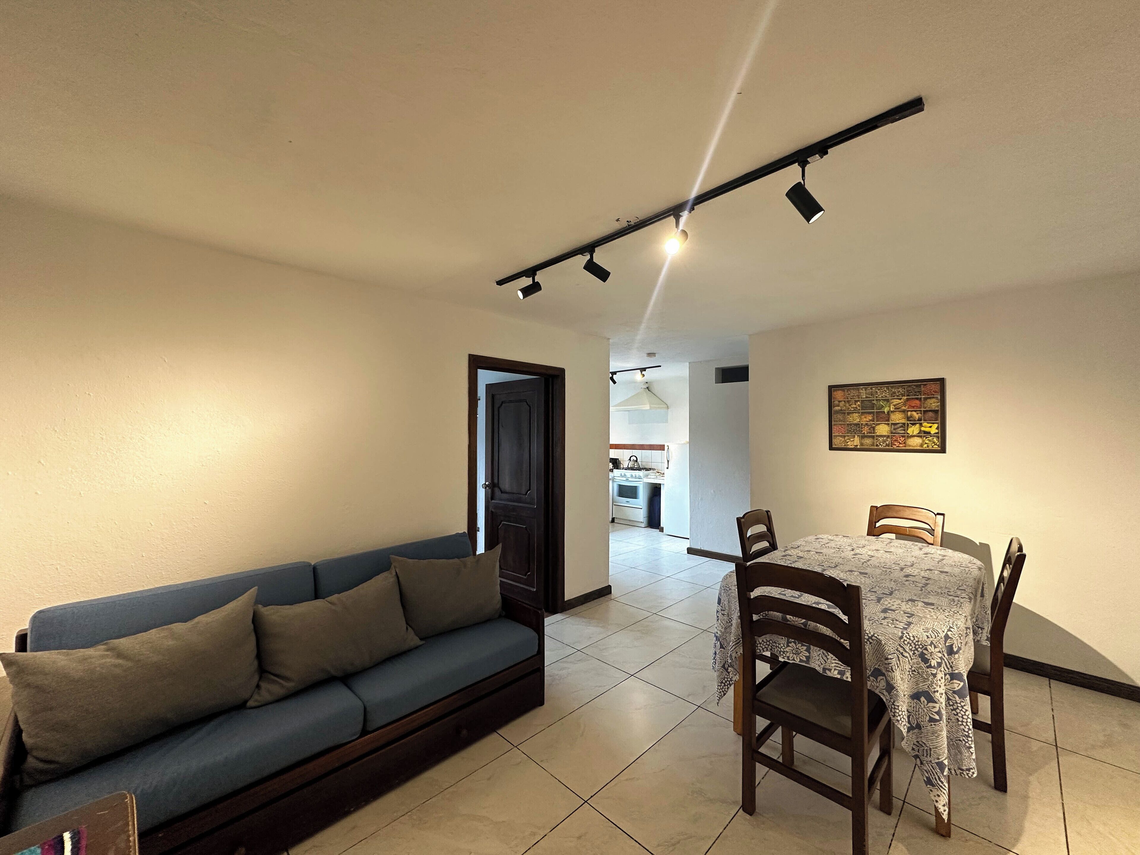 A living room with a sofa, a dining table with chairs, and a kitchen in the background.