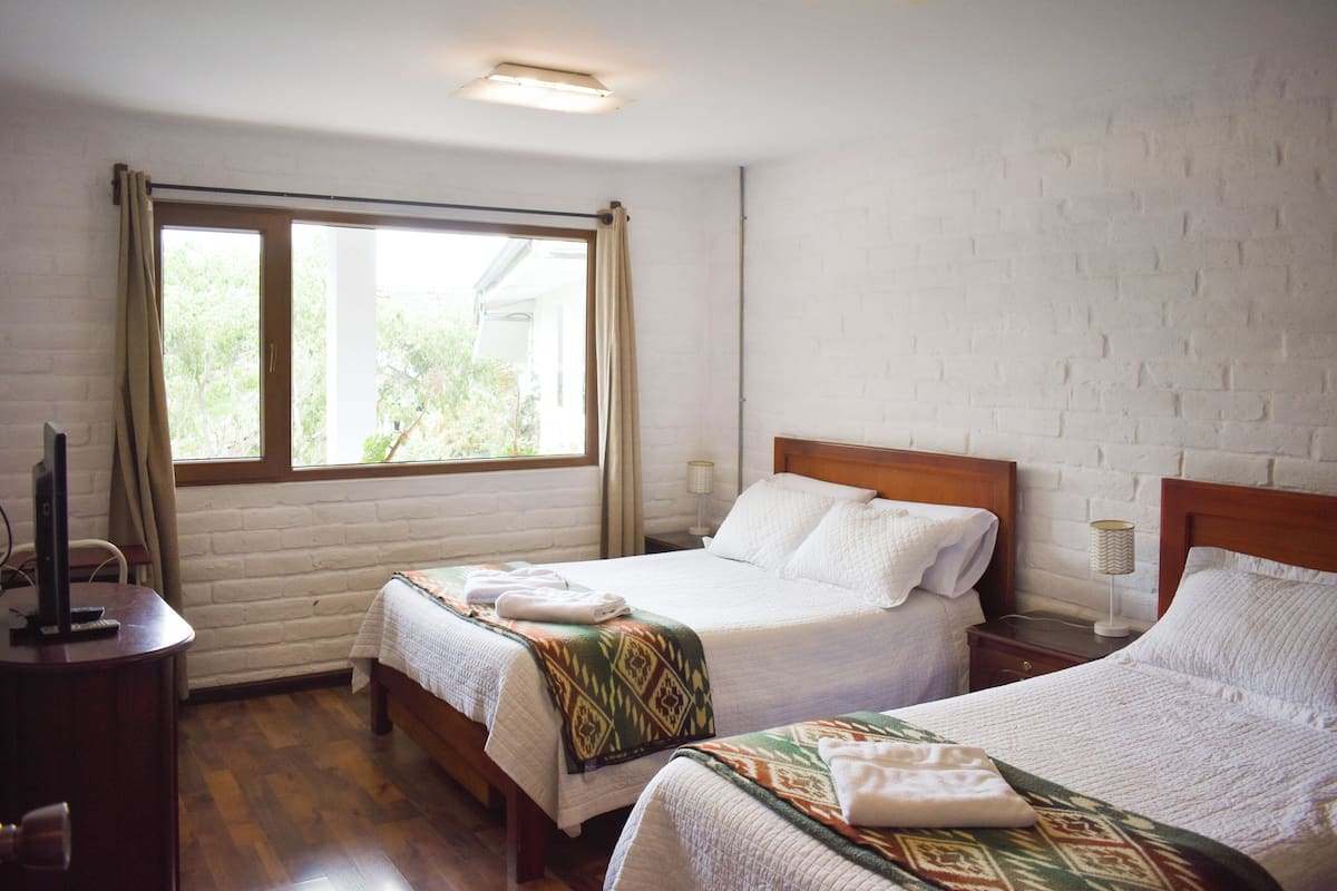 Habitación de hotel con dos camas, suelo de madera, pared de ladrillo y ventana con cortinas.