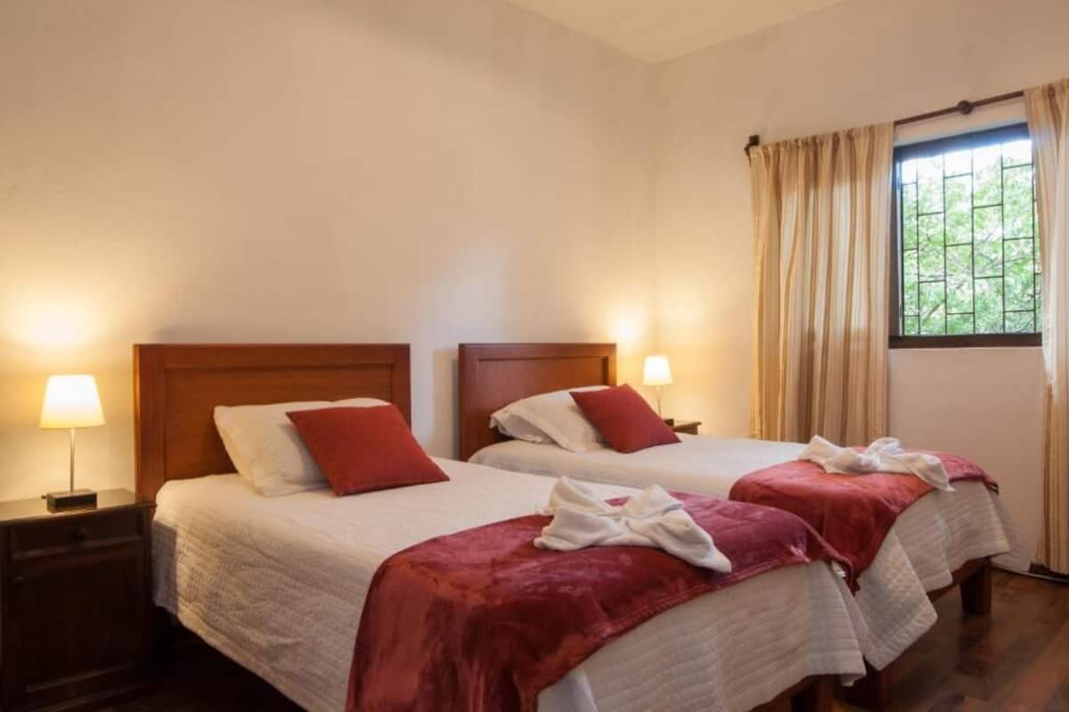 Habitación de hotel con dos camas, cabeceros de madera, lámparas de noche y una ventana con cortinas.