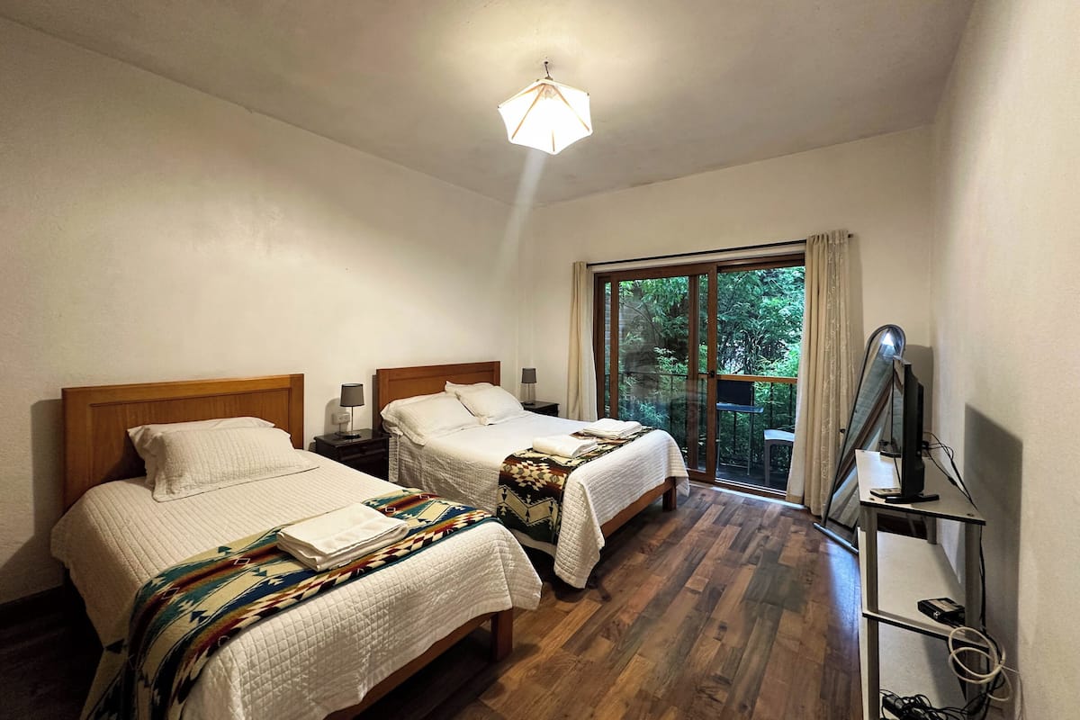 Habitación de hotel con dos camas, un amplio ventanal corredero a un balcón, un escritorio con computadora e iluminación en el techo.