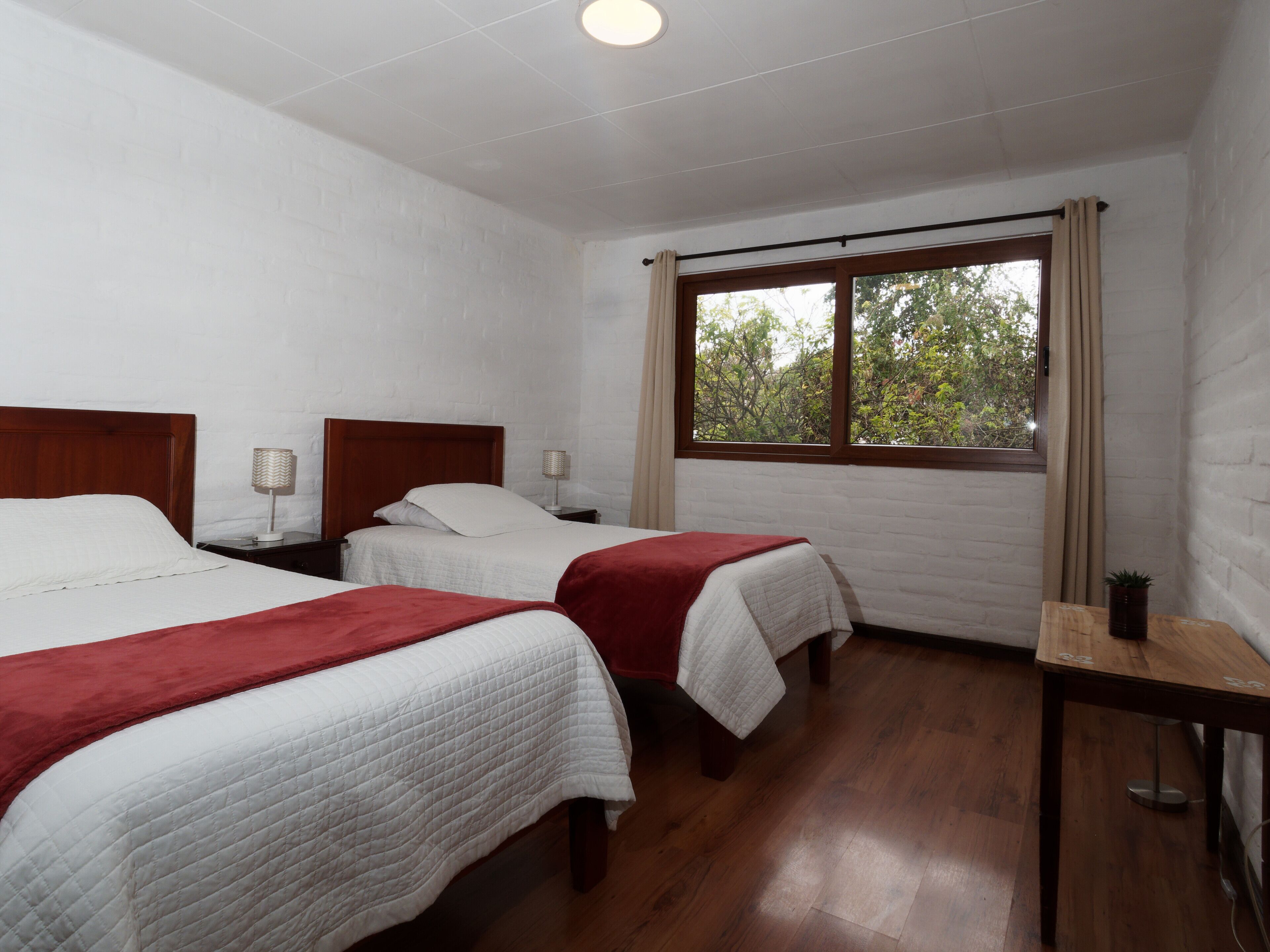 A hotel room with two beds, wooden flooring, white brick walls, a window with curtains, and a small table with a lamp.