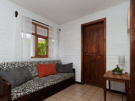 Una habitación con un sofá estampado, una puerta de madera, una mesita con una planta y una ventana con cortinas de gasa.