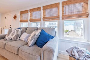 A living room with a grey sofa, multiple pillows, and a view through large windows with wooden blinds. - The Granite Harbor (Wells)