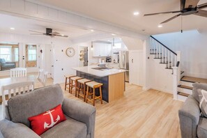 A spacious living area with a grey sofa, wooden stools, and a red pillow. A kitchen with white cabinets and a central island is visible. - The Granite Harbor (Wells)