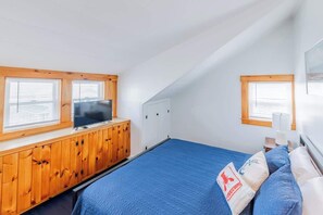 A bedroom with a sloped ceiling, a bed with blue bedding, a wooden cabinet, a television, and a window with blinds.