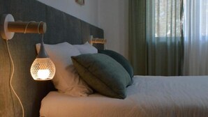 A neatly made bed with a headboard, a bedside lamp, and a window with curtains. - Old Village Hostel (Ponte de Lima)