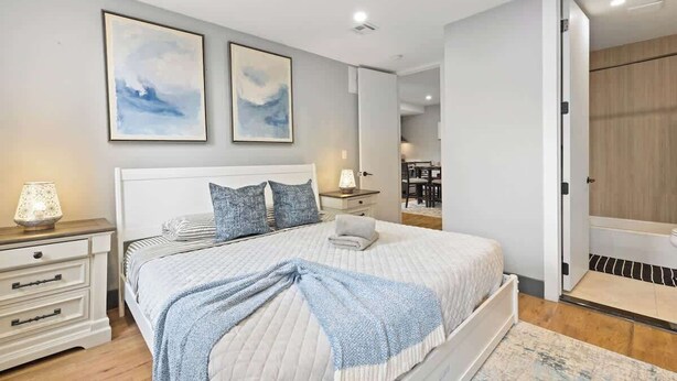 A bedroom with a bed, bedside tables, and a nightstand. There are two framed pictures on the wall, a lamp on the nightstand, and a bathroom visible through an open door.
