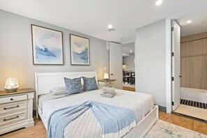 A bedroom with a bed, bedside tables, and a nightstand. There are two framed pictures on the wall, a lamp on the nightstand, and a bathroom visible through an open door.