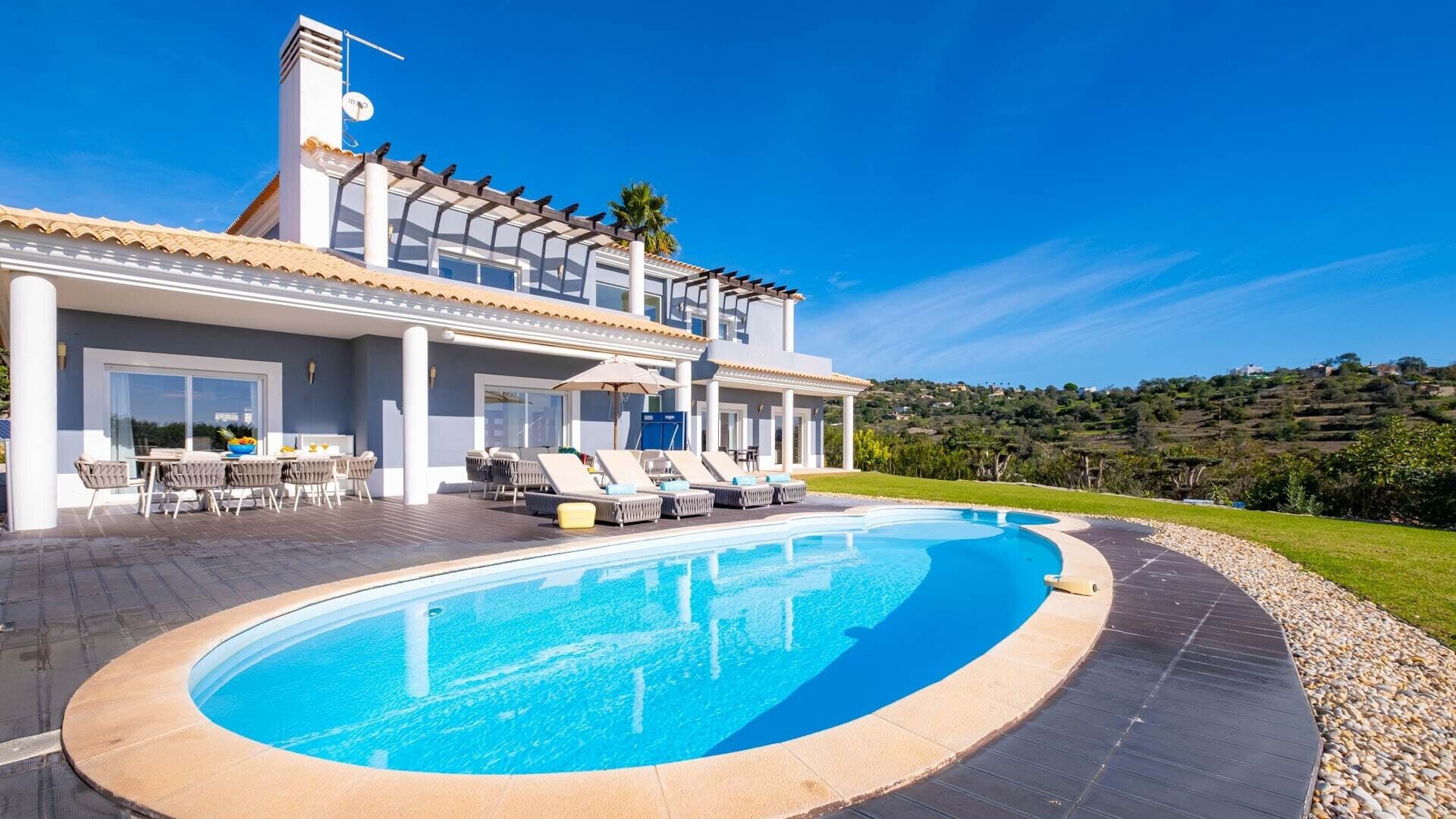 A modern villa with a large swimming pool, outdoor seating, and a scenic hillside view.