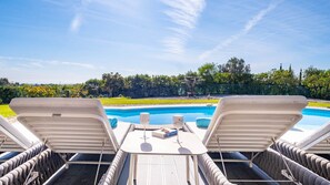 A poolside lounge with two sun loungers facing a pool, a table with a glass and a book, and a view of the pool and greenery.