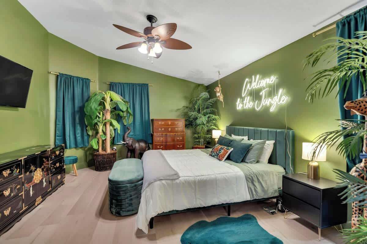 A bedroom with a bed, a ceiling fan, a neon sign, and various plants.