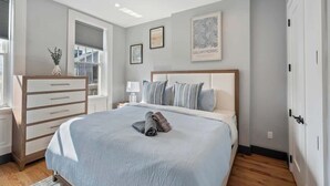 Um quarto com cama, penteadeira, mesa de cabeceira e janela com persianas.
